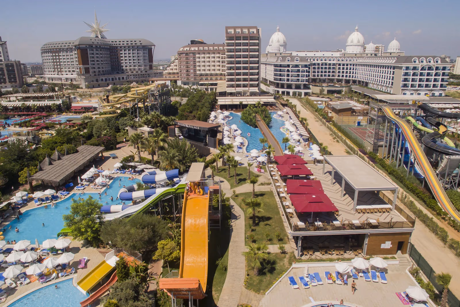 Luchtfoto van de zwembaden, de gele en blauwe glijbanen en het terras met rode parasols bij Saturn Palace Resort in Lara