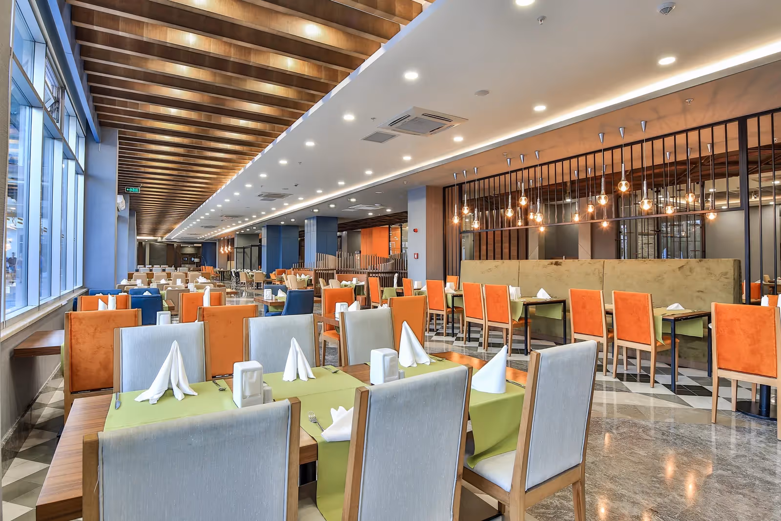 Lange restaurantzaal met gedekte tafels, oranje stoelen en grote ramen in Saturn Palace Resort in Lara