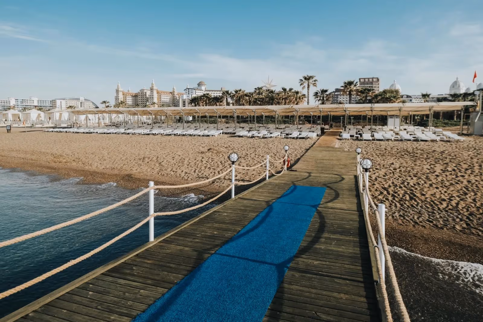 Houten pier met touwreling over het water bij Saturn Palace Resort, met het strand en de ligbedden erachter