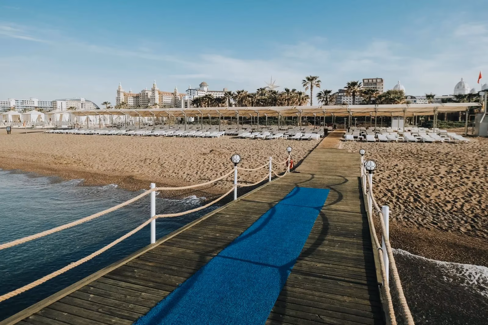 Houten pier met touwreling over het water bij Saturn Palace Resort, met het strand en de ligbedden erachter