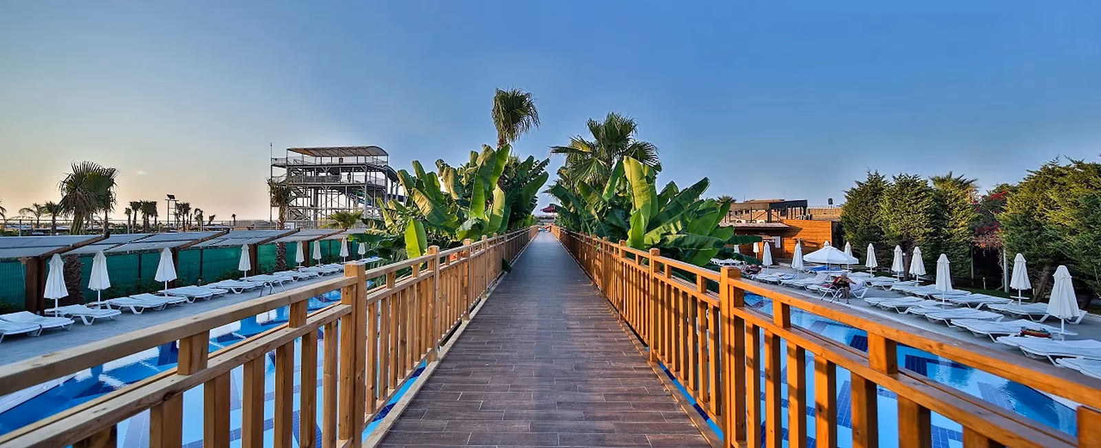 Houten wandelpad met reling over het zwembad, met palmen en ligbedden aan weerszijden bij Saturn Palace Resort