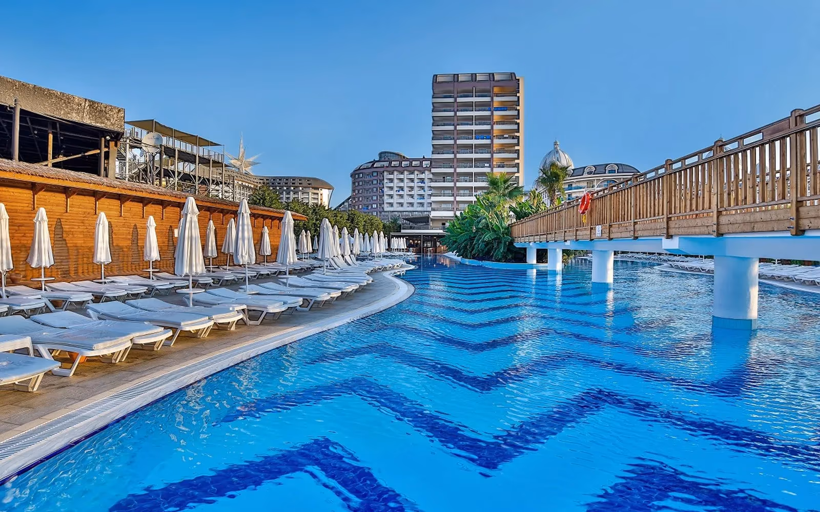 Zwembad met blauw zigzagpatroon, ligbedden en een houten brug, met de hoteltoren op de achtergrond bij Saturn Palace Resort in Lara