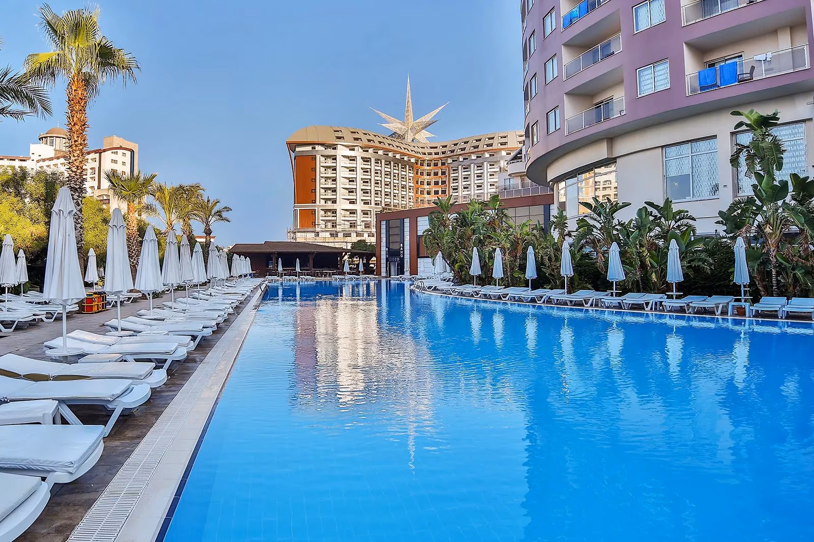 Lang rechthoekig zwembad met rijen ligbedden en parasols tussen de hotelgebouwen van Saturn Palace Resort