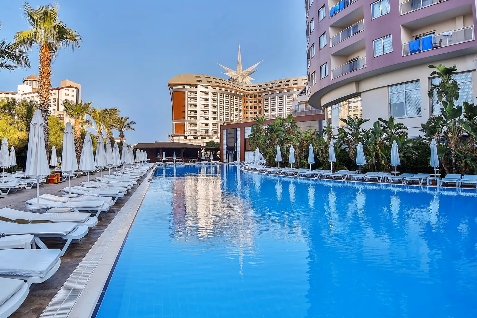 Lang rechthoekig zwembad met rijen ligbedden en parasols tussen de hotelgebouwen bij Saturn Palace Resort in Lara