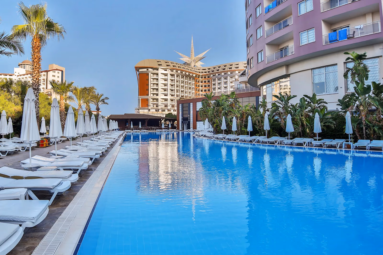 Lang rechthoekig zwembad met rijen ligbedden en parasols tussen de hotelgebouwen bij Saturn Palace Resort in Lara